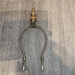 May include: A brass lamp harp featuring a decorative finial at the top. The harp consists of a U-shaped metal frame with two vertical attachment points at the base, designed to support a lampshade on a table or floor lamp fixture.