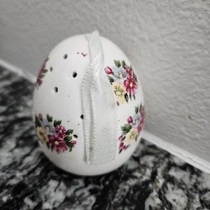 以下が含まれることがあります： ピンク、黄色、青の花柄が施された、白いセラミック製の卵型の塩入れ。塩入れの上部にはいくつかの小さな穴と白いリボンが付いています。装飾的なキッチンアイテムです。