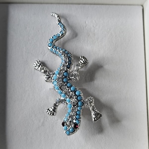 May include: A silver-toned lizard brooch with a turquoise and clear crystal design. The lizard has a curved tail, four legs, and two red eyes. The brooch is set against a white background.