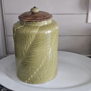 Może przedstawiać: Jasnozielony ceramiczny słoik z wzorem liści, zwieńczony brązową drewnianą pokrywką i małym zielonym guzikiem. Słoik jest cylindryczny i stoi na białym owalnym talerzu.