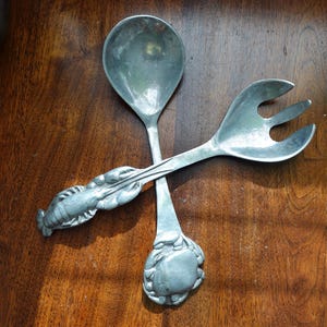 Puede incluir: Un juego de dos utensilios de servicio de plata con diseños de langosta y cangrejo. Los utensilios están sobre una superficie de madera.