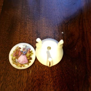 May include: Two-piece porcelain set. One piece is a small, round, white lid with a painted scene of a couple in period clothing. The other piece is a white, decorative object with a small spout and two curved handles.