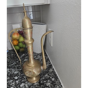 Könnte beinhalten: Eine verzierte Messingkanne mit langem, gebogenem Ausguss und Griff. Die Kanne hat aufwendige geätzte Designs und einen dekorativen Deckel. Die Farbe ist ein warmes Gold mit roten und schwarzen Akzenten. Die Kanne wird wahrscheinlich zum Servieren von Getränken verwendet.