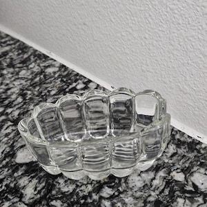 Op de afbeelding: Een heldere glazen schaal met een geschulpte rand. De schaal heeft een rechthoekige vorm met afgeronde hoeken en een gestructureerd oppervlak. Het glas is transparant, waardoor licht erdoorheen kan. Het wordt waarschijnlijk gebruikt om te serveren of tentoon te stellen.