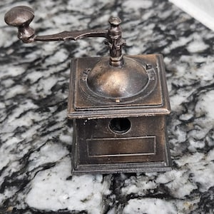 May include: A vintage-style, bronze-colored pencil sharpener with a hand crank. The sharpener has a rectangular base with a circular opening and a small, decorative handle. The crank arm extends from the top, and the overall design is reminiscent of antique coffee grinders.