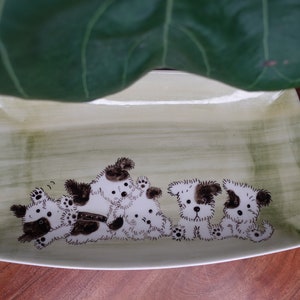 Puede incluir: Un plato rectangular de cerámica con un esmalte verde y blanco. El plato presenta un diseño pintado a mano de cinco perros juguetones en blanco y negro.