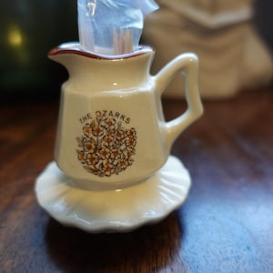 May include: White ceramic pitcher with a brown rim and a floral design. The pitcher says "The Ozarks" on the front. It is sitting on a white ceramic base.