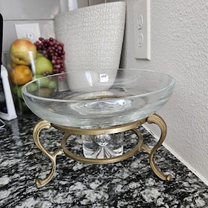 Op de afbeelding: Een heldere glazen kom met een brede rand staat op een goudkleurige metalen standaard. De standaard heeft gebogen poten en een cirkelvormige basis. De kom is leeg en de achtergrond bevat een vaas en een bak met fruit.