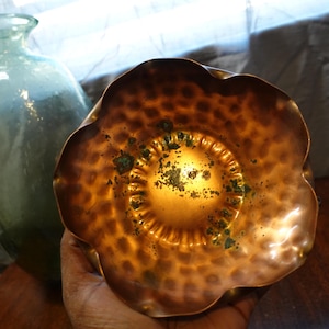 May include: A copper flower-shaped bowl with a hammered texture and a green patina. The bowl has a scalloped edge and a slightly raised center.