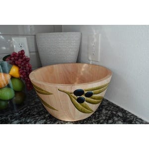 May include: A wooden bowl with a light finish, featuring a painted olive branch design with green leaves and black olives. The bowl is empty and sits on a dark countertop. The bowl is likely used for serving or display.