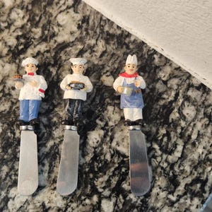 Puede incluir: Tres cuchillos de mantequilla decorativos con figuras de chef. Cada cuchillo presenta una figura de chef diferente, vestida con uniformes blancos y azules, sosteniendo alimentos. Los cuchillos tienen hojas plateadas y están colocados sobre una encimera moteada.