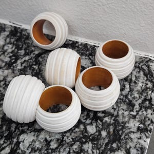 May include: Six white and beige striped napkin rings with a wooden interior. The rings are round with a grooved exterior design. These decorative rings are perfect for adding a touch of elegance to any table setting.