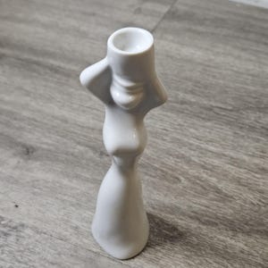 May include: A white ceramic candle holder shaped like a stylized human figure with arms raised to support the circular candle cup at the top. The smooth, glossy finish covers the entire abstract form, which features a narrow waist and a flared base.