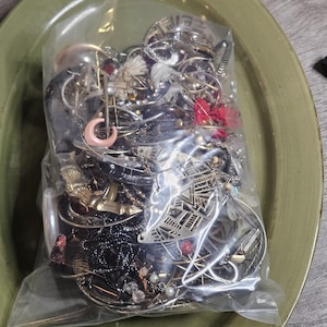 May include: A clear plastic bag overflowing with assorted jewellery items: rings, earrings, necklaces, and bracelets. The pieces are in silver, gold, and black, with red and pink details. The bag rests on a green oval plate, offering a glimpse of the collection.