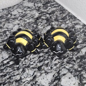 Puede incluir: Dos figuras de arañas de cerámica con cuerpos negros y abdómenes a rayas amarillas y negras. Las arañas tienen múltiples patas y caras sonrientes. Las figuras están sobre una superficie gris moteada.