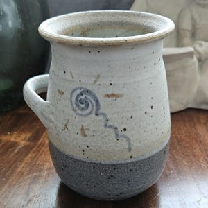 May include: A white ceramic mug with a speckled glaze and a hand-painted sun design. The mug has a handle and a gray base.
