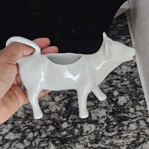 May include: A white ceramic cow creamer with a handle and a spout. The creamer is in the shape of a cow, with four legs and a raised head. The creamer is designed to hold liquid and pour it out.