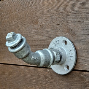 May include: A silver metal pipe hook with a round flange. The hook is attached to a wooden wall.