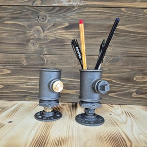 Op de afbeelding: Twee zwarte metalen pijp potloodhouders met messing accenten. De houders staan op een houten oppervlak en elk heeft een ander assortiment aan pennen en potloden.