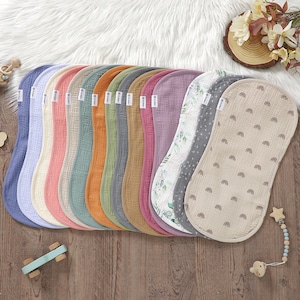 Puede incluir: Una colección de baberos para bebés en varios colores, incluyendo azul, rosa, naranja y verde. Algunos baberos presentan patrones como arcoíris y diseños florales. Los baberos están hechos de una tela suave y texturizada y están dispuestos sobre una superficie de madera.