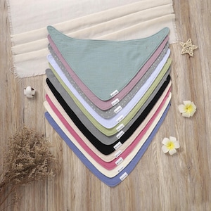 Op de afbeelding: Een stapel van tien baby slabbetjes in verschillende kleuren, waaronder blauw, roze, zwart en groen. Elk slabbetje is driehoekig, gemaakt van een zachte, getextureerde stof en heeft een klein label. Ze zijn gerangschikt op een houten oppervlak met decoratieve elementen.