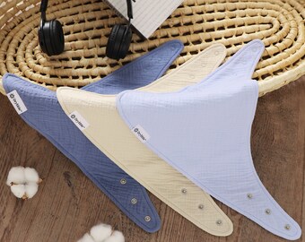 Puede incluir: Cuatro baberos triangulares para bebés en tonos azules y crema, con cierres de botón a presión. Los baberos están hechos de una tela texturizada y están colocados sobre una superficie de madera. La marca "Drybble" es visible.