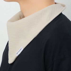 May include: A beige bib-style scarf with a textured surface and a contrasting light beige trim. The scarf is worn over a black shirt. A small white tag with the text "Drybib" is visible on the scarf.