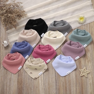 May include: A collection of baby bibs in various colors, including pink, blue, and beige. Some bibs have a solid color, while others feature a rainbow pattern. The bibs are made of soft, textured fabric and are arranged on a wooden surface.