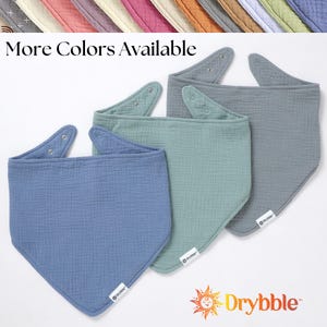 May include: Three cotton baby bibs in different colors: blue, green, and gray. The bibs are triangular shaped with a rounded bottom and have a small logo on the bottom corner. The text "Drybbble" is printed in orange and yellow on the bottom of the image.