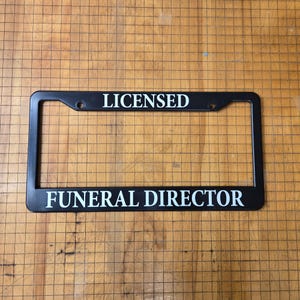May include: Black plastic license plate frame with white text that reads "Licensed Funeral Director".