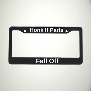 May include: Black license plate frame with the text "Honk If Parts" above and "Fall Off" below in white lettering. The frame has a rectangular shape with rounded corners and a matte finish.