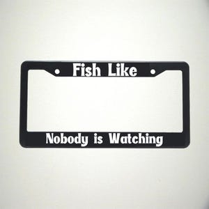 May include: Black license plate frame with the text "Fish Like" at the top and "Nobody is Watching" at the bottom in white lettering. The frame has a rectangular shape with rounded corners and two small holes.