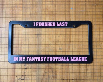 May include: Black license plate frame with the text "I finished last in my fantasy football league" in pink lettering.