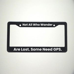 May include: Black license plate frame with white text that reads "Not All Who Wander Are Lost. Some Need GPS." The frame has a rectangular shape with rounded corners and a matte finish.
