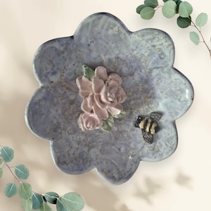 May include: A ceramic decorative plate with a flower-shaped design. The plate is a marbled blue-gray color with a cluster of pink roses and a bee figurine. The plate has a scalloped edge and is a unique home decor item.
