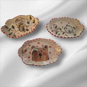 May include: Three small, oval-shaped ceramic trinket dishes with scalloped edges and red-dotted rims. One dish features a brown bear face with red hearts, another a bird on a branch, and the third a leafy branch design, each holding metal rings.