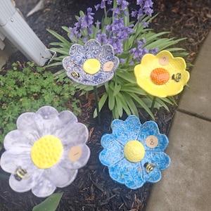 May include: Four ceramic flower-shaped garden decorations in shades of blue, yellow, and lavender. Each flower has a yellow center and a small bee figurine. The flowers are placed in a garden bed with mulch and greenery.
