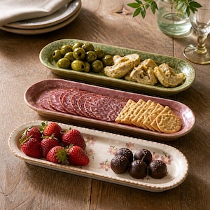 May include: Three elongated oval ceramic serving platters arranged vertically. The top platter is sage green with a textured rim, holding green olives and crackers. The middle platter is dusty rose with a textured rim, holding salami and crackers. The bottom platter is cream with a floral pattern, holding fresh strawberries and dark chocolate truffles.
