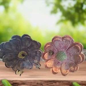 May include: Two decorative flower-shaped objects. One is dark purple with a yellow center and a small bee. The other is pink with a green center and a small bee. Both are on a wooden surface.