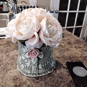 May include: A ceramic vase with a green and white patterned design, featuring a pink flower accent. The vase is filled with a bouquet of pale pink silk peonies.
