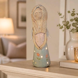 May include: A tall, slender ceramic angel figurine with long, textured blonde hair, a pale pink bodice featuring subtle white polka dots, and a light blue skirt decorated with small orange flowers. The base of the skirt has cutouts emitting light.
