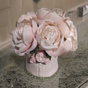 May include: A white ceramic vase with a woven pattern, filled with pink silk flowers. The vase is sitting on a gray countertop.