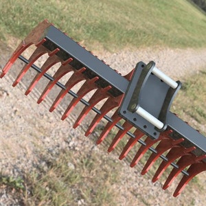 Könnte beinhalten: Ein roter Metallrechenaufsatz für einen Traktor. Der Rechen hat einen schwarzen Metallrahmen und einen Griff mit zwei weißen Metallstangen.