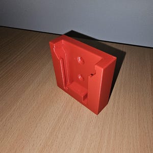 May include: A 3D printed red plastic wall mount with a cutout for a tool. The mount has two holes for screws.