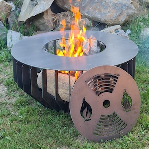 Puede incluir: Un pozo de fuego de metal negro con una parte superior redonda y una placa de metal decorativa con un diseño de llama. El pozo de fuego está rodeado de rocas y hierba verde.