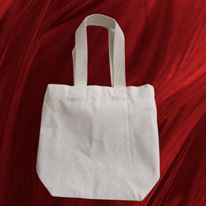 May include: A small, off-white canvas tote bag with sturdy handles. The bag is rectangular with a slightly rounded bottom and is set against a red background.