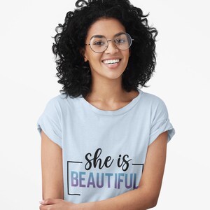 May include: A light blue t-shirt with the text "she is BEAUTIFUL" in black and purple. The person in the image is wearing round glasses and has curly black hair. The person is smiling.