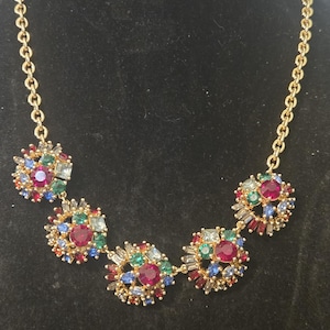 May include: A gold-toned necklace featuring five floral-shaped pendants. Each pendant is adorned with a cluster of colorful gemstones in shades of red, blue, green, and clear. The necklace has a delicate chain.