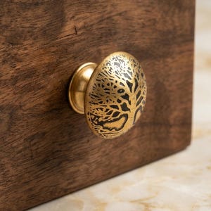 Può includere: Un pomello rotondo per mobile in ottone con un design ad albero inciso in scuro sulla parte frontale. Il pomello è montato su una superficie in legno marrone scuro con venature visibili, caratterizzato da una finitura metallica con dettagli neri.