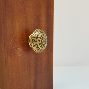 May include: A round cabinet knob featuring a gold and black geometric mandala pattern, mounted on a smooth, medium-brown wooden surface. The knob has a circular centre design surrounded by repeating floral-like motifs in a metallic finish.